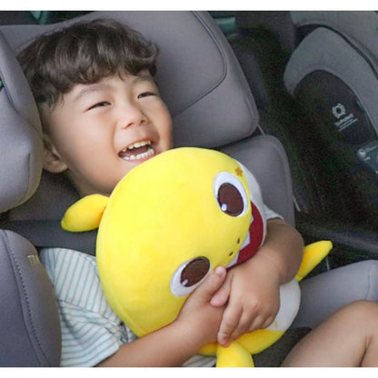 一個開心的小男孩坐在汽車安全座椅上，懷裡緊緊抱著一個黃色的 Baby Shark 鯊魚寶寶安全帶抱枕，笑得非常燦爛。