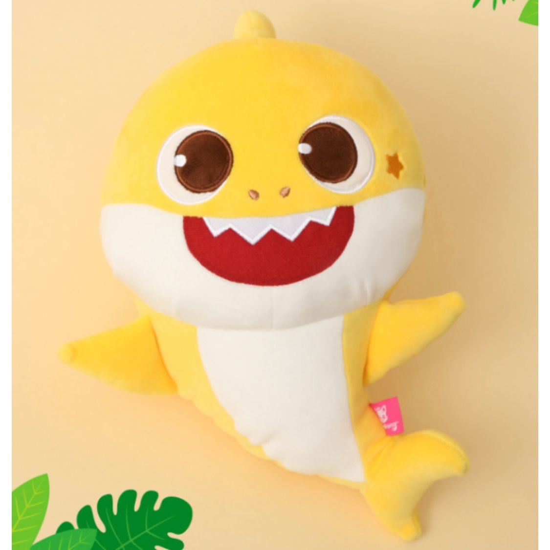 Pinkfong 正版授權 Baby Shark 黃色鯊魚寶寶絨毛玩偶特寫，擁有大大的眼睛、紅色的嘴巴與頭上的星星標誌，背景為淡黃色並有綠色葉子裝飾。
