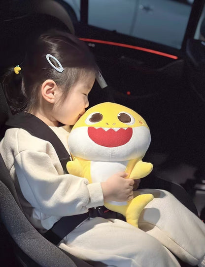 一個開心的小女孩坐在汽車安全座椅上，懷裡緊緊抱著一個黃色的 Baby Shark 鯊魚寶寶安全帶抱枕，笑得非常燦爛。