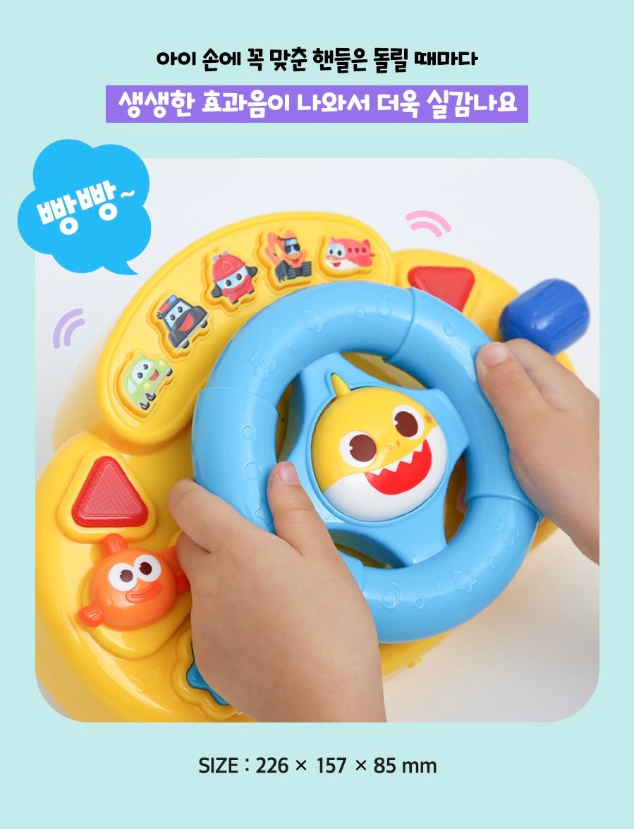一個幼兒的雙手握著黃色底座、藍色握把的 Baby Shark 玩具方向盤。方向盤中央有立體的鯊魚寶寶臉部造型，上方有多個交通工具圖案按鈕。圖片文字顯示「叭叭～」的對話框。